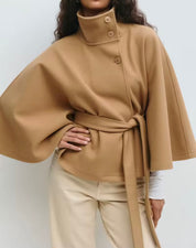High-Neck Belted Cape Jacket in camel wool blend. Poncho-inspired autumn/winter coat with high-neck collar, cape silhouette and adjustable waist belt. A chic belted jacket combining elegance, comfort and versatile seasonal style.