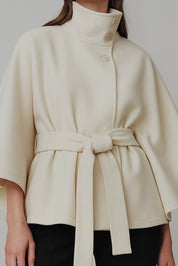 High-Neck Belted Cape Jacket in camel wool blend. Poncho-inspired autumn/winter coat with high-neck collar, cape silhouette and adjustable waist belt. A chic belted jacket combining elegance, comfort and versatile seasonal style.