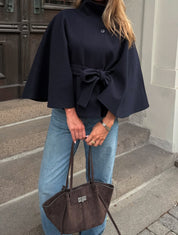 High-Neck Belted Cape Jacket in camel wool blend. Poncho-inspired autumn/winter coat with high-neck collar, cape silhouette and adjustable waist belt. A chic belted jacket combining elegance, comfort and versatile seasonal style.
