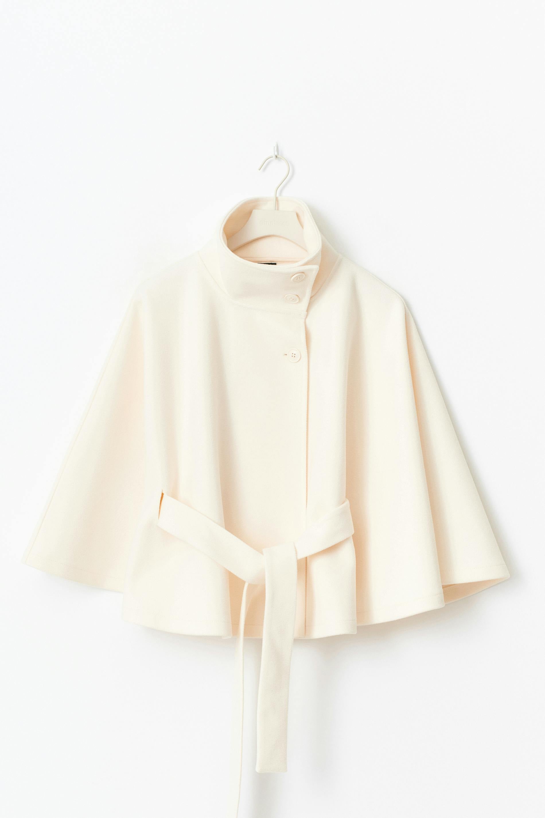 High-Neck Belted Cape Jacket in camel wool blend. Poncho-inspired autumn/winter coat with high-neck collar, cape silhouette and adjustable waist belt. A chic belted jacket combining elegance, comfort and versatile seasonal style.
